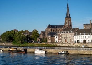 Discover Maastricht's charming riverside with historic buildings and the iconic church under a bright summer sky.